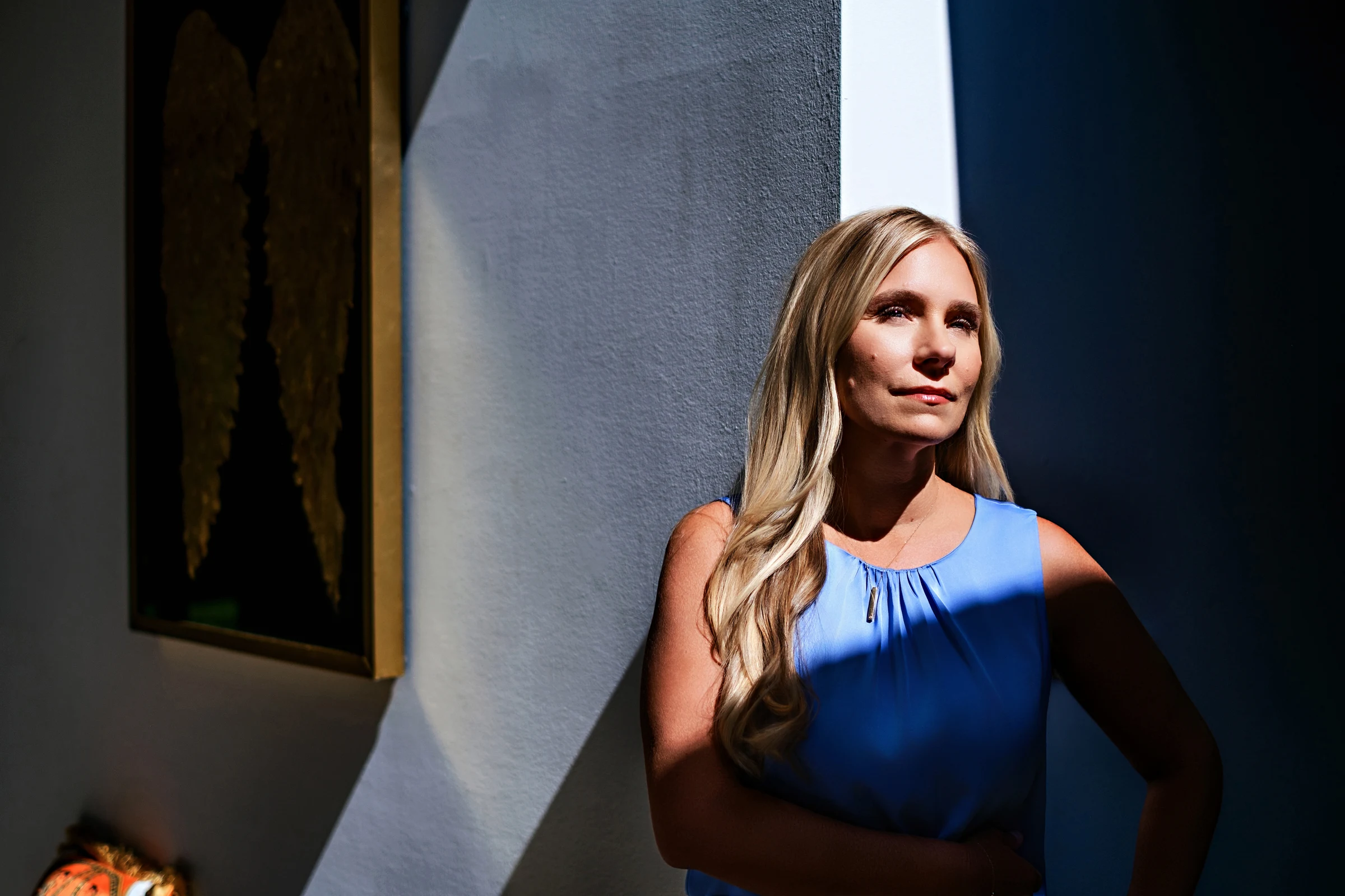 Brittany Conner in a blue sleeveless top, standing in diagonal sunlight beside framed artwork